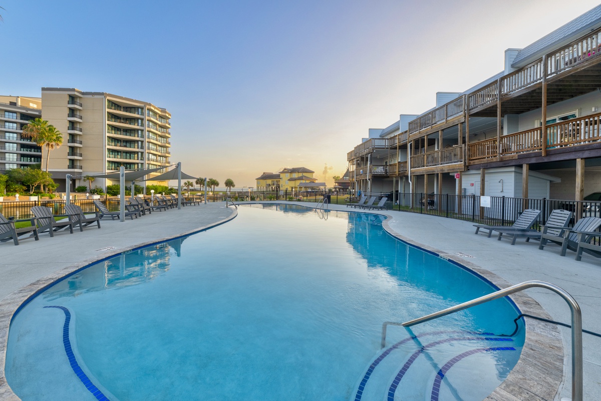 A spacious curved pool with ample loungers invites you to soak up the sun, surrounded by multi-level balconies and coastal charm under golden skies.