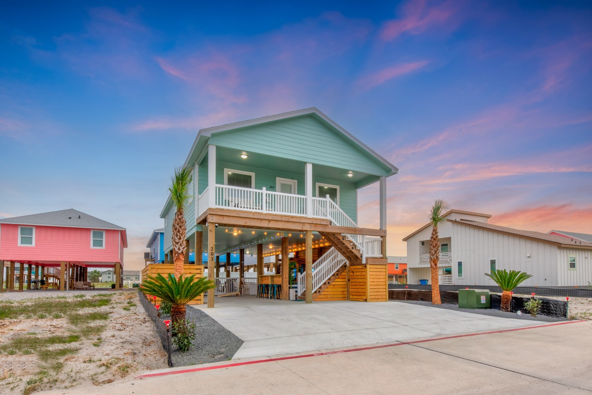 A charming green beach house on stilts with tropical landscaping and outdoor entertaining space, set against a stunning sunset sky.