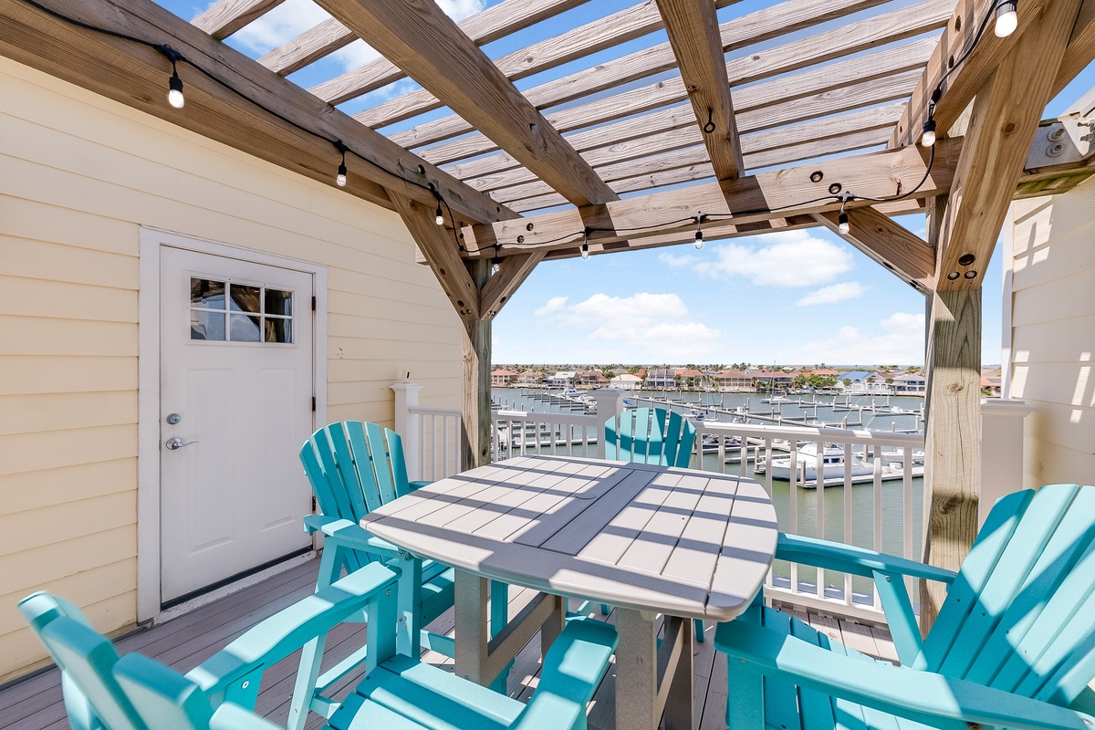 Relax on your private waterfront deck with turquoise Adirondack chairs overlooking the marina, where pergola shade and string lights create perfect ambiance.