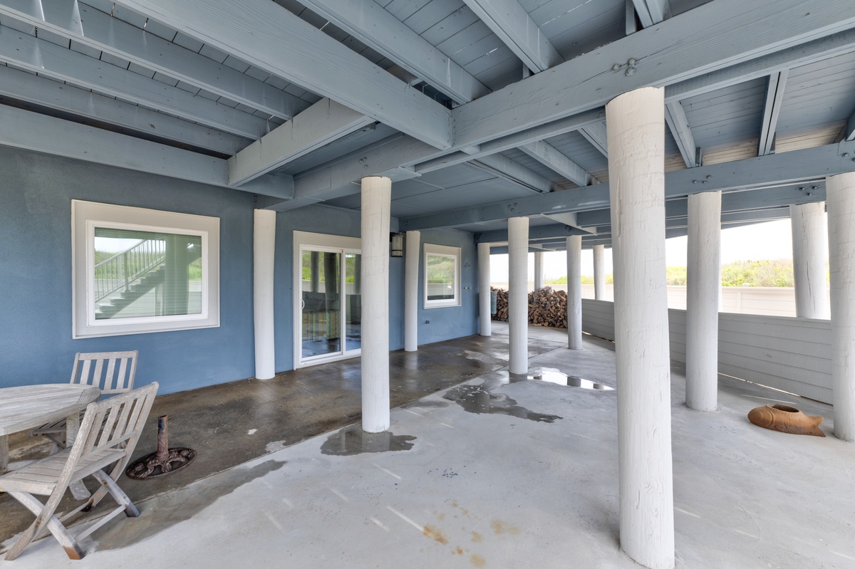 Covered ground-level area with concrete flooring, white support columns, and blue exterior walls featuring windows and outdoor seating.