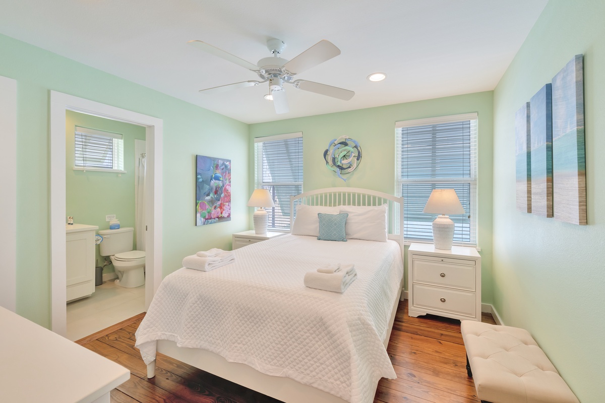 Relax in your serene mint-green bedroom with coastal charm, featuring a comfy white bed, ceiling fan, and private bathroom.
