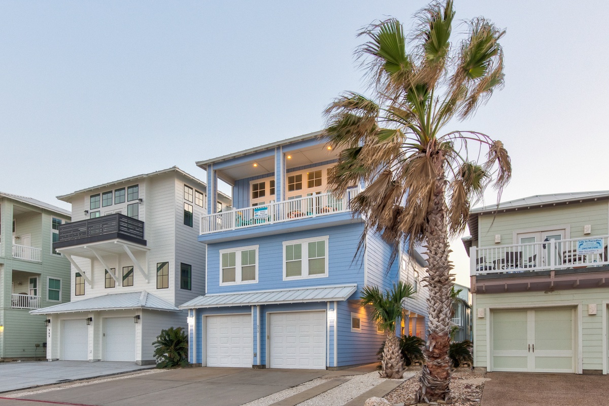 Bright blue beachside home with palm trees and multiple balconies in a coastal neighborhood setting.