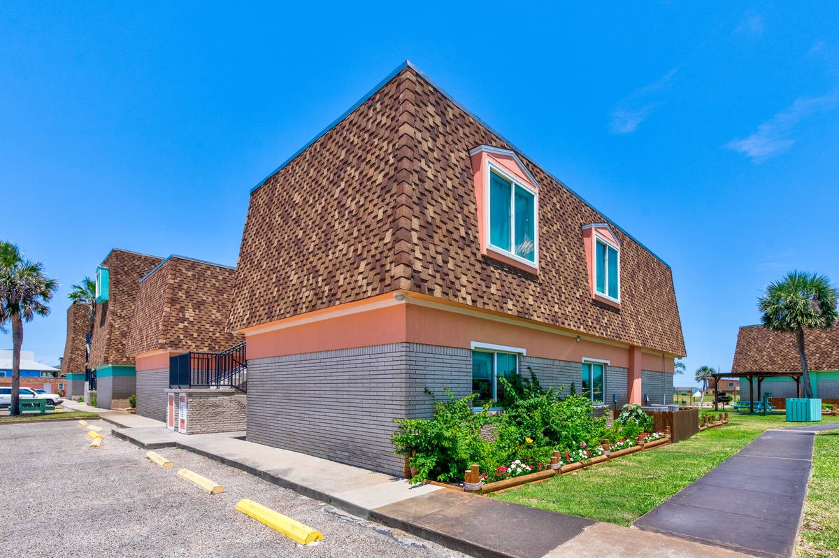 Modern vacation rental with colorful coral exterior and distinctive shingled roof, surrounded by tropical landscaping in a peaceful coastal neighborhood.