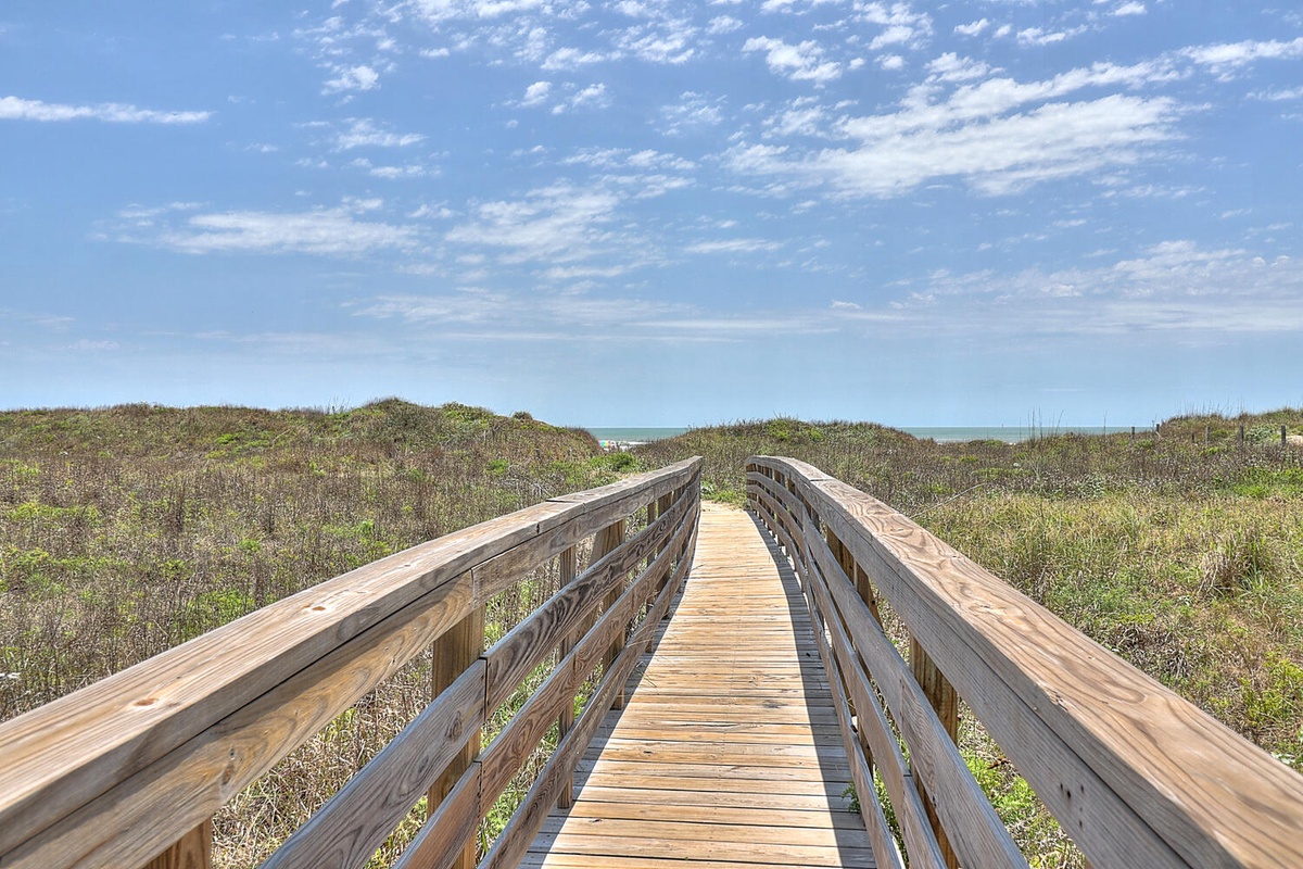 Wooden boardwalk winds through coastal dunes under a vast blue sky dotted with white clouds.