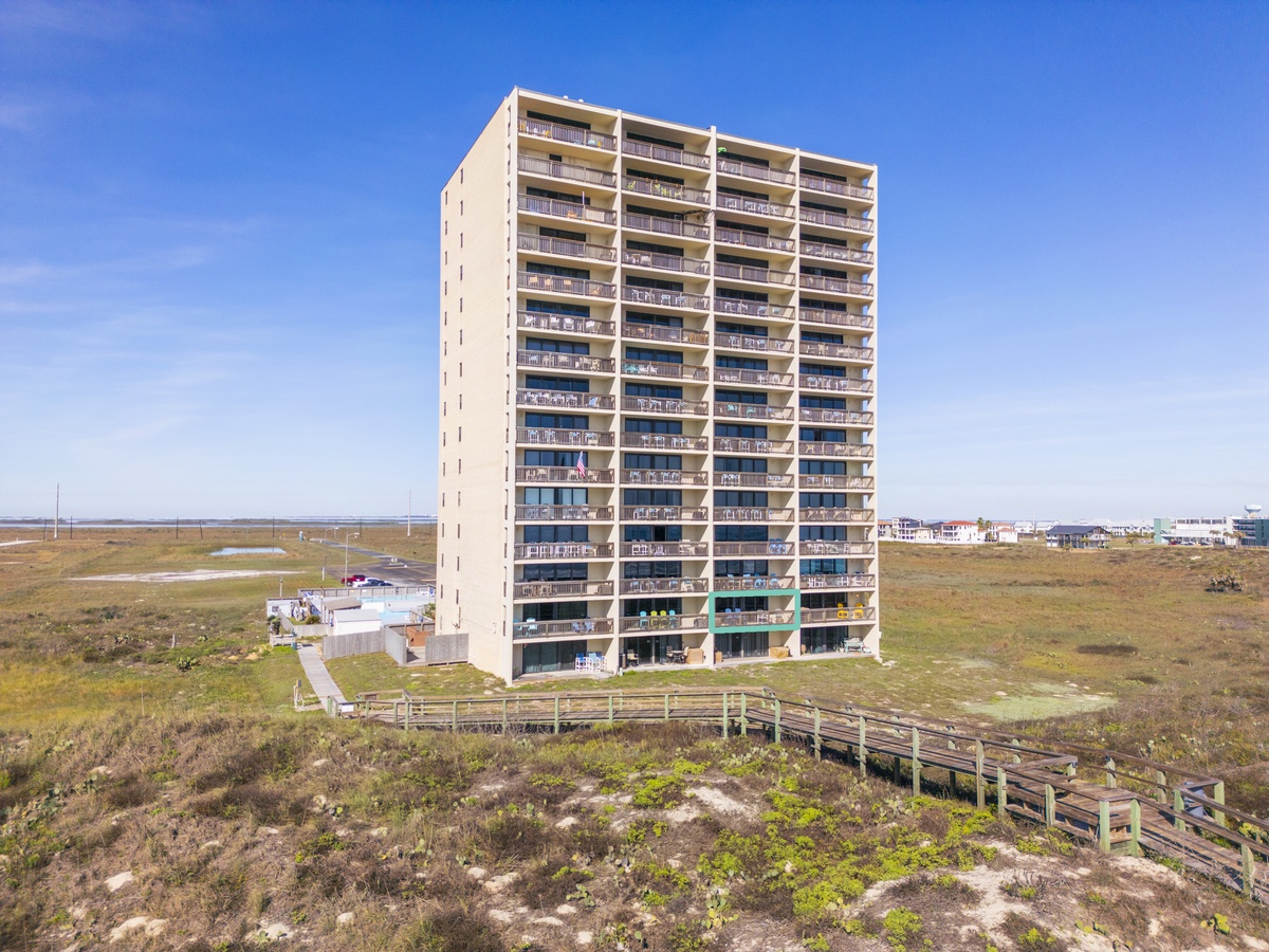 Beachfront high-rise building set among coastal dunes with wooden boardwalk access under clear blue skies.