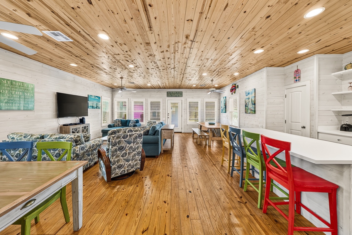 Step into your spacious, light-filled living area where rustic wood ceilings and gleaming hardwood floors set a warm, welcoming tone. Cozy seating, colorful bar stools, and plenty of natural light make this your perfect gathering spot.