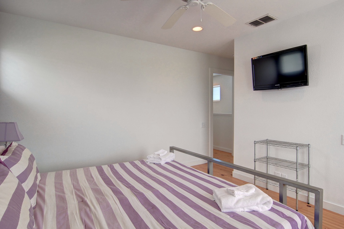 Unwind in your peaceful bedroom with purple-striped bedding, ceiling fan, and wall-mounted TV for cozy evenings.