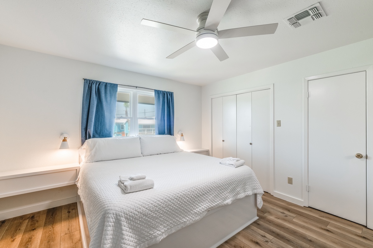 Drift off in your bright, comfortable bedroom with soft linens, gentle fan breeze, and charming blue curtains framing peaceful views.