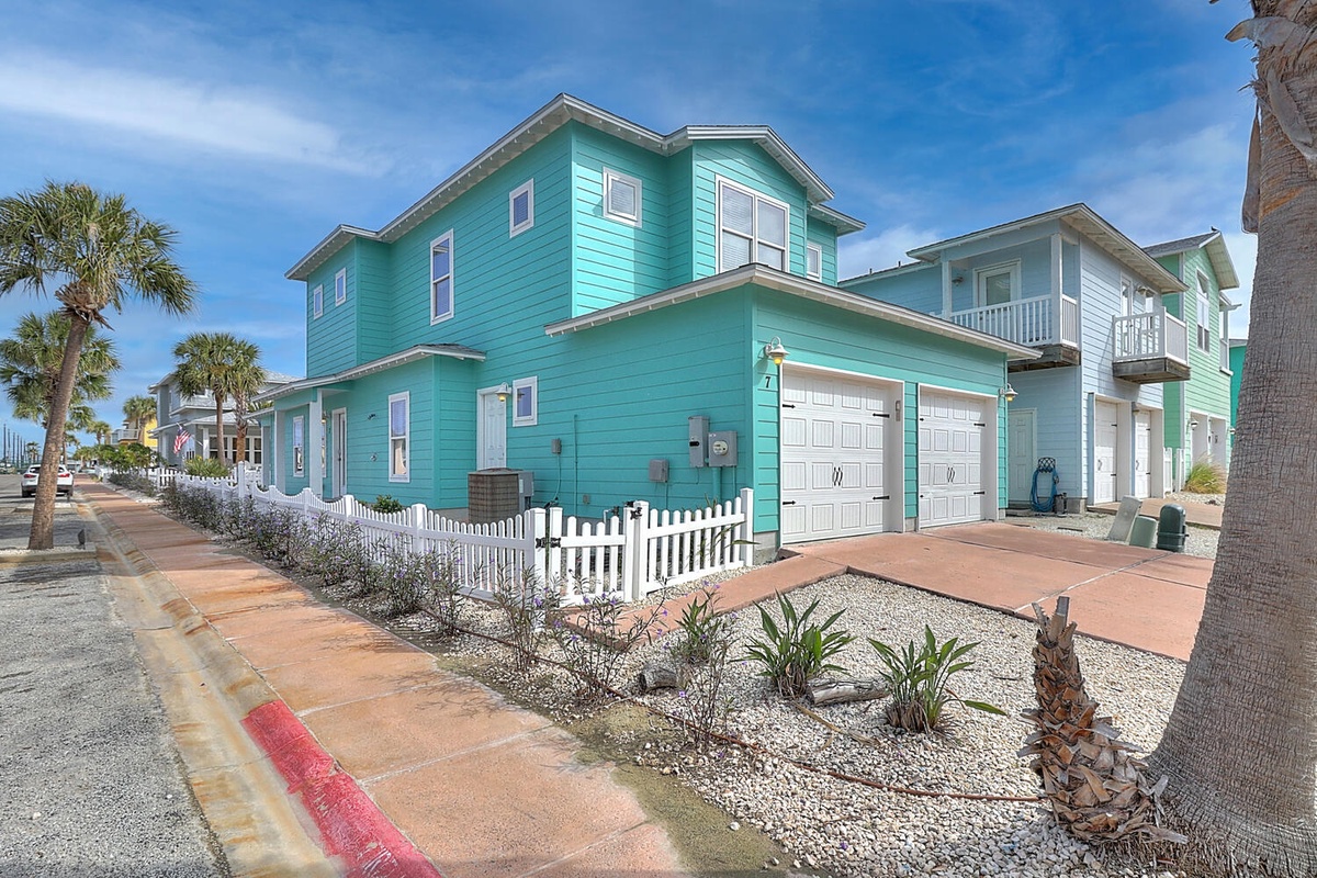 Charming turquoise beach house with white picket fence and tropical landscaping in a vibrant coastal neighborhood.