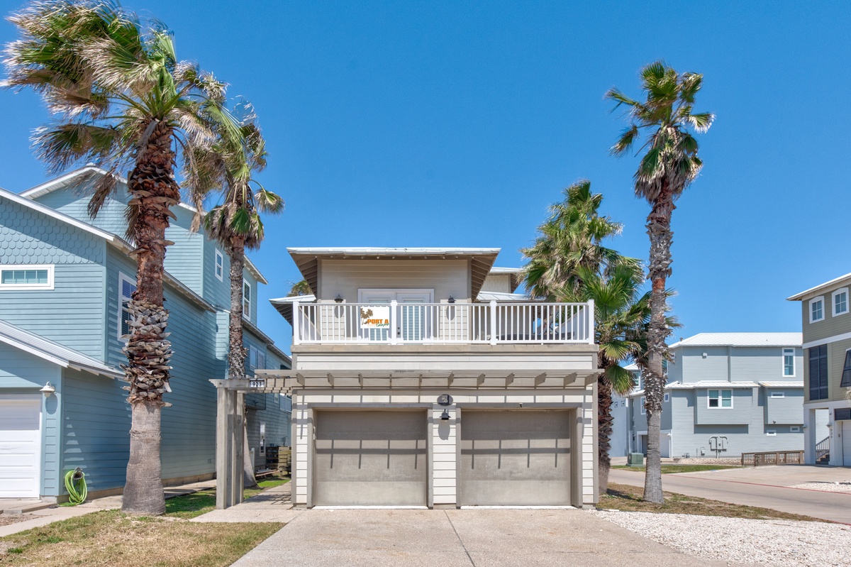 Modern coastal property featuring two-car garage and upper-level balcony, surrounded by swaying palm trees in a residential beach community.
