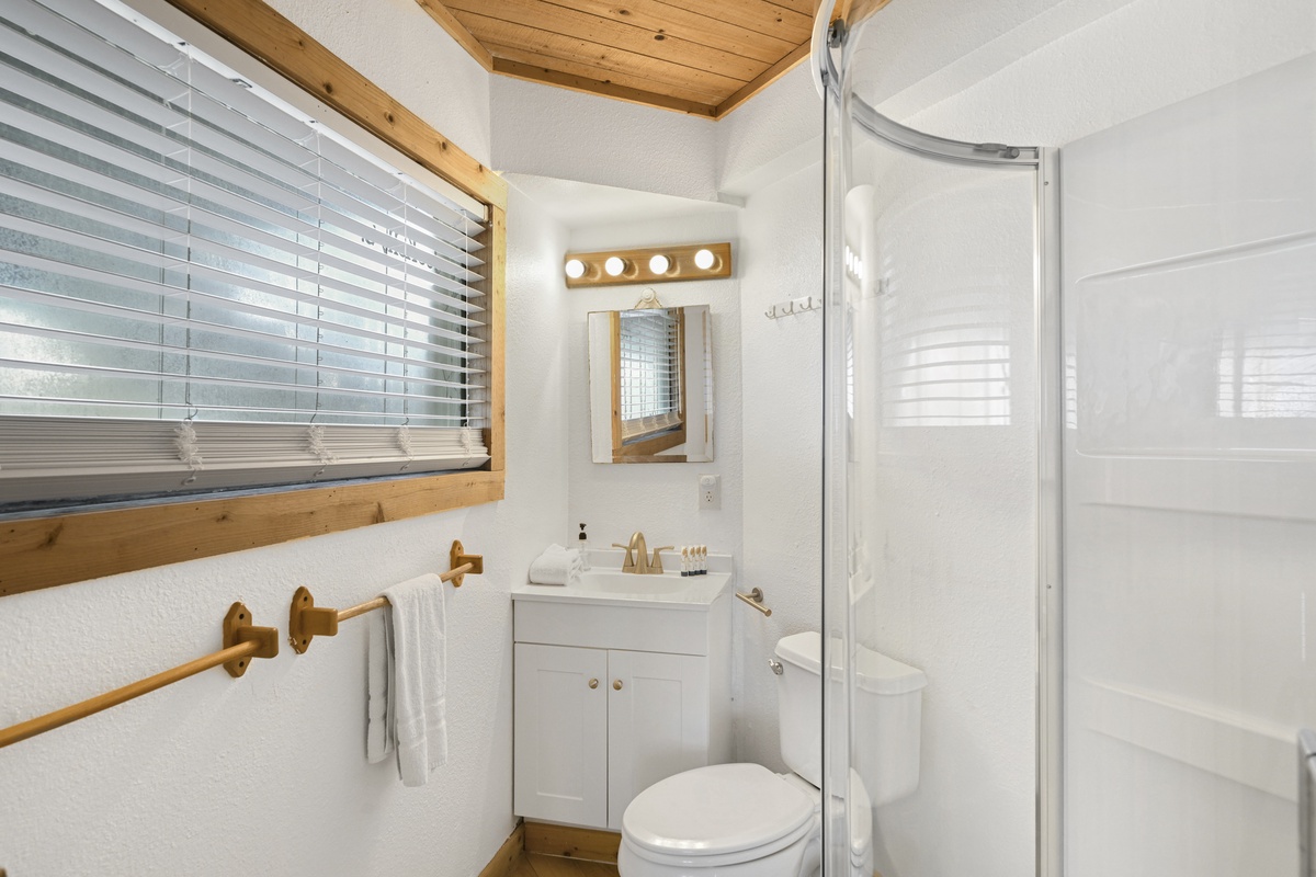 Step into a bright, thoughtfully designed bathroom with warm wood accents and modern fixtures. Sunlight filters through the window, highlighting crisp white walls, a spacious shower, and all the comforts you need to refresh and unwind.