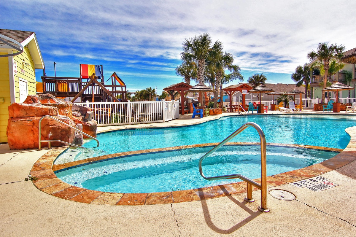 Vibrant resort-style pool with rock waterfall and children's playground nearby, surrounded by colorful beach cottages and swaying palm trees.