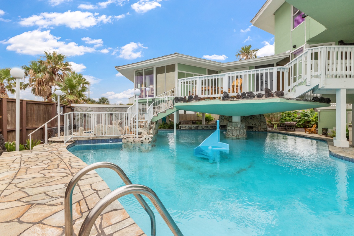 Dive into your private paradise with a stunning pool featuring a thrilling water slide and tropical landscaping under endless blue skies.