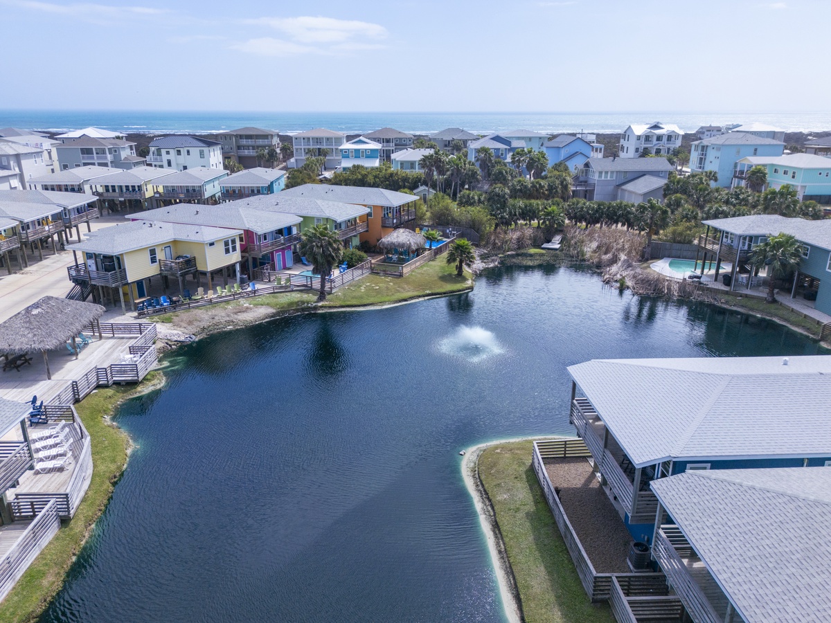 Aerial view of a coastal community featuring colorful beach homes surrounding a serene pond, with the ocean visible in the distance under a bright sunny sky.