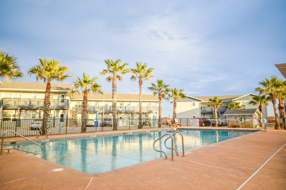 Pool area surrounded by palm trees and multi-story resort buildings under clear blue skies during daytime.