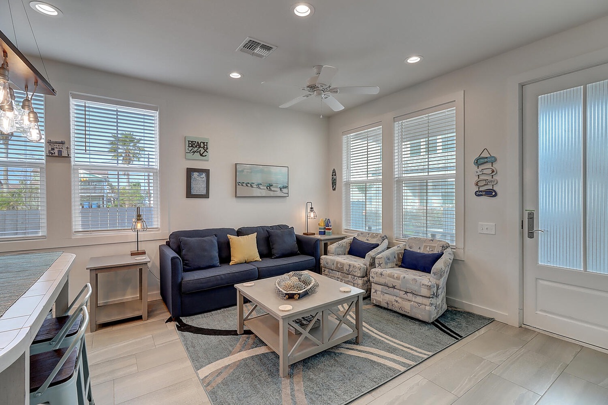 Relax in this bright, coastal-themed living room with comfortable seating, nautical artwork, and plenty of natural light streaming through blinds.