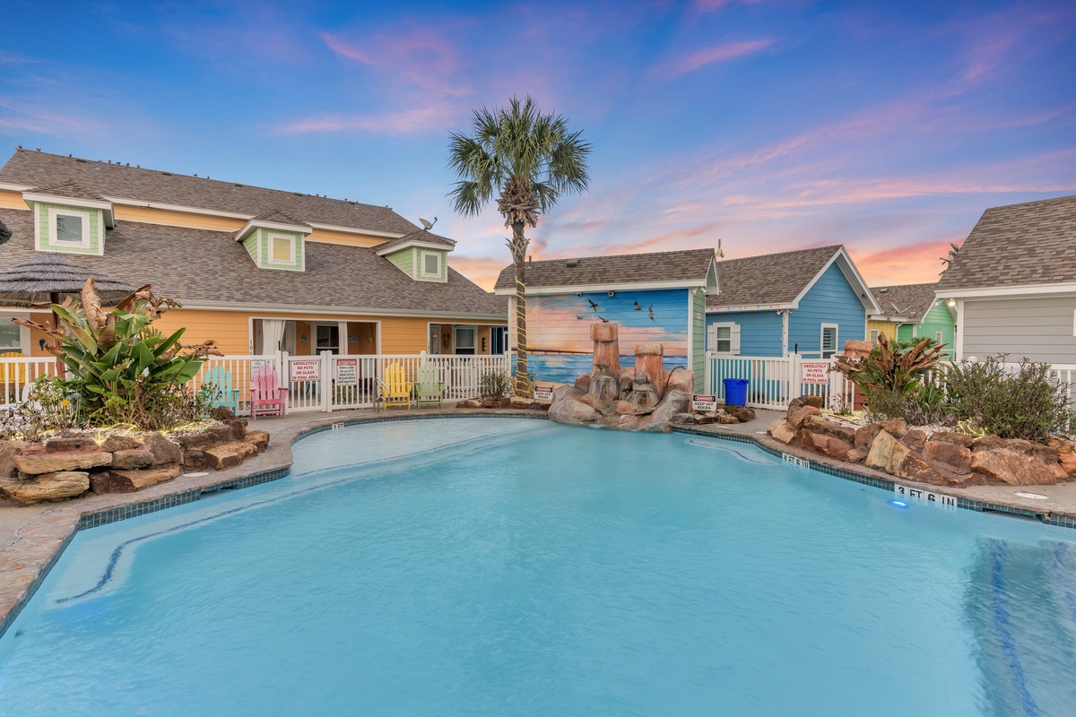 Colorful beach-themed cottages surround a sparkling pool area with tropical landscaping and stunning sunset skies overhead.