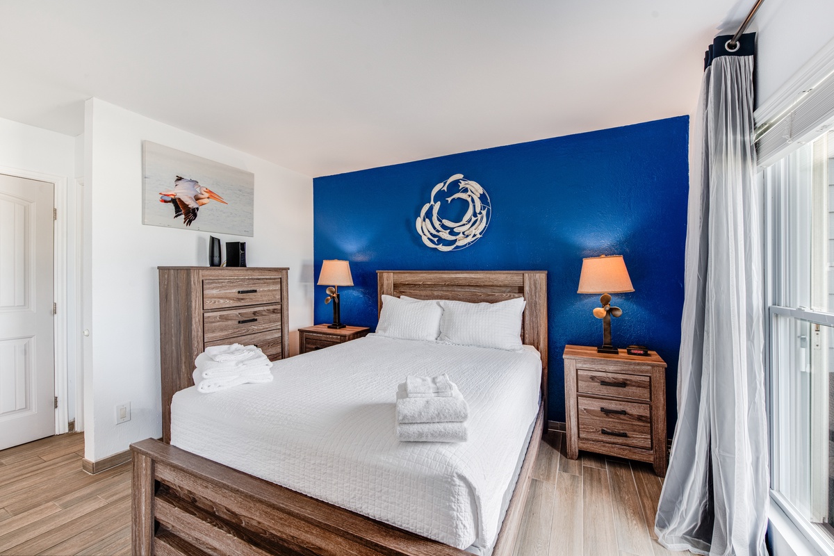 Sink into crisp white linens against a stunning ocean-blue accent wall, where warm wood tones and thoughtful touches create your perfect retreat.