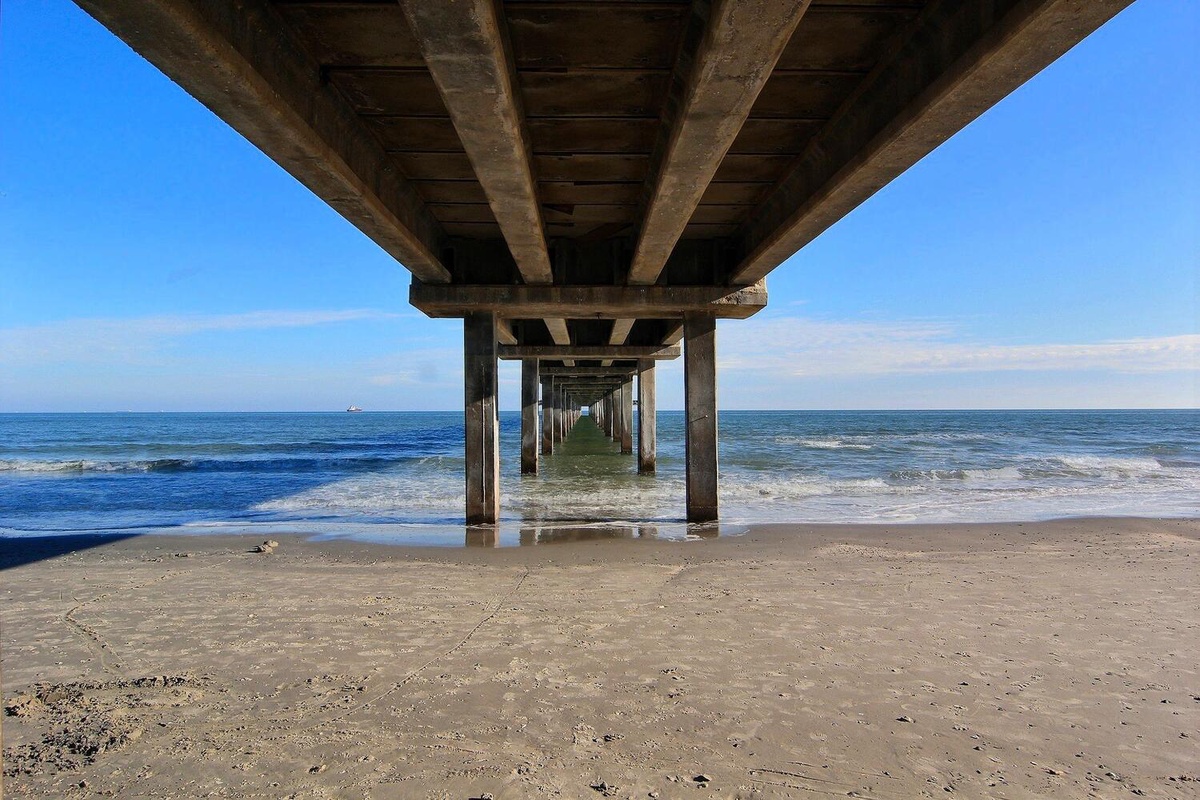 A weathered pier extends over sandy beach and rolling waves under bright blue skies.