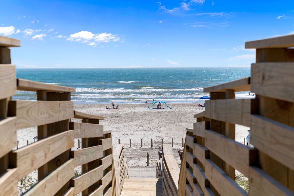 Wooden boardwalk leads down to pristine sandy beach with ocean waves and scattered beachgoers under bright blue skies.