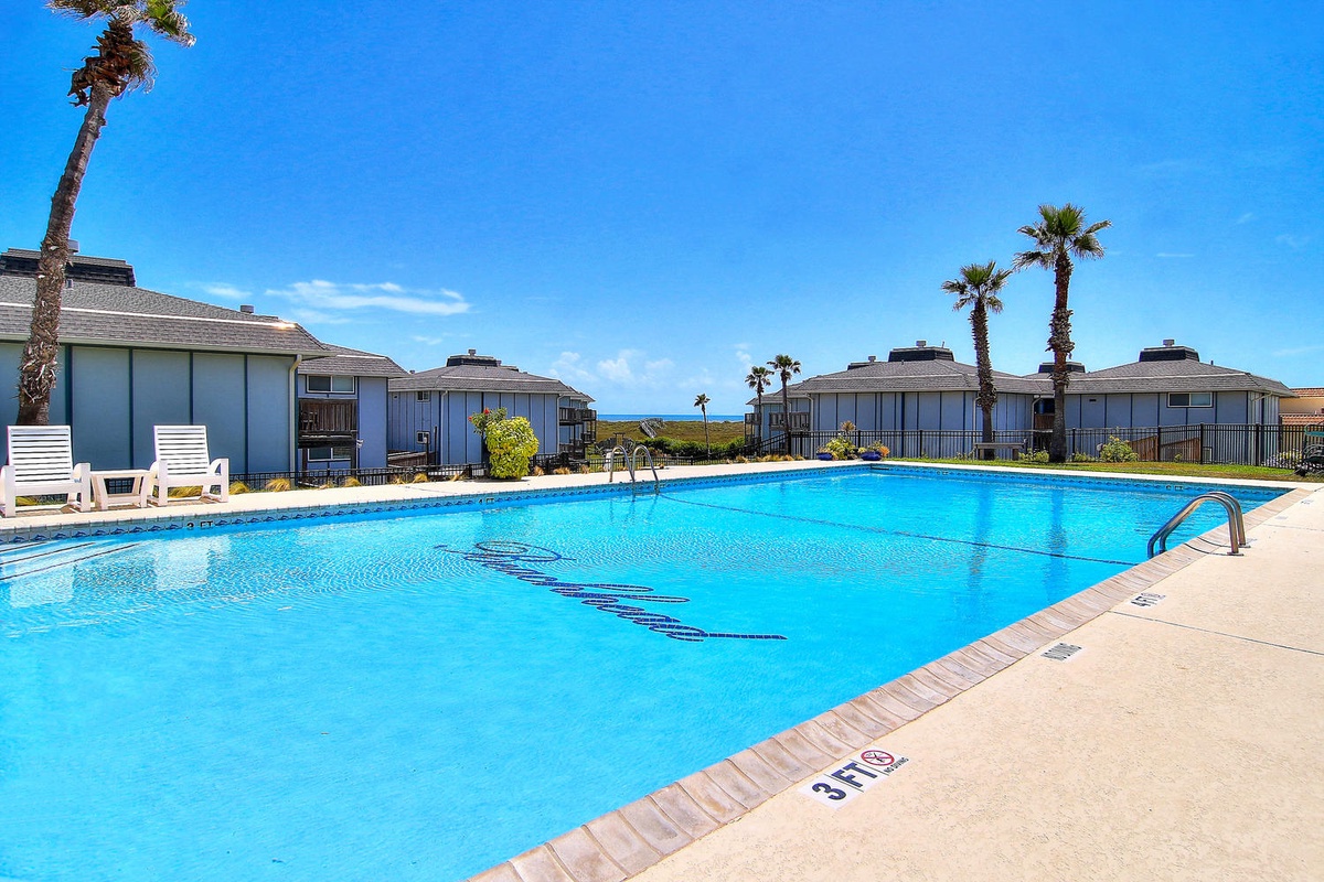 Sparkling community pool with ocean glimpses beyond palm-lined units under perfect blue skies.