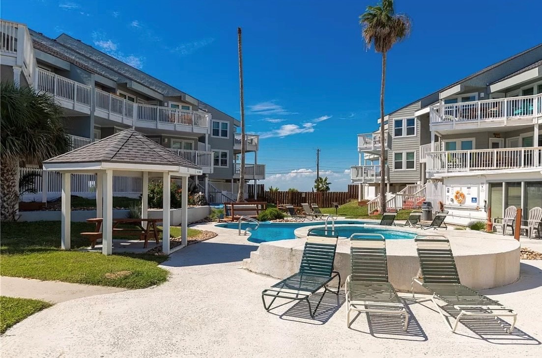 Spacious vacation rental community featuring a sparkling pool area with loungers and covered gazebo, surrounded by multi-level buildings with balconies.