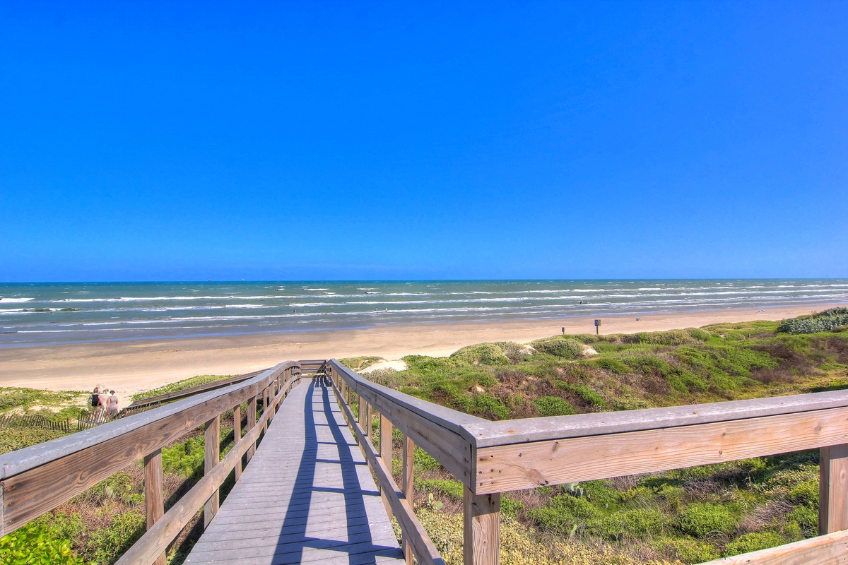 Wooden boardwalk leads to pristine sandy beach with rolling waves and endless blue sky.