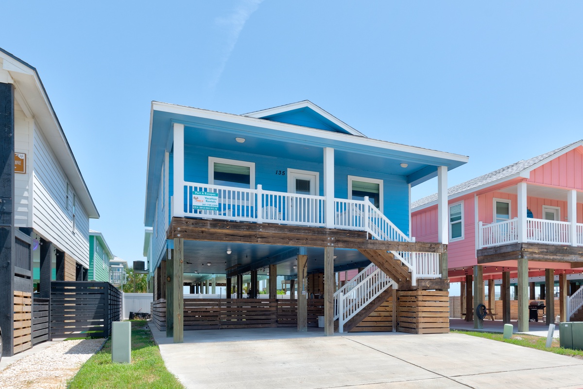 This bright coastal home sits elevated on sturdy pilings in a welcoming neighborhood, with covered parking below and a cheerful blue exterior accented by white railings and columns.