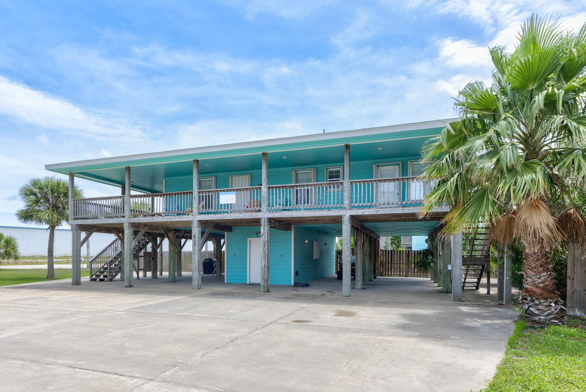 Coastal vacation home elevated on stilts with turquoise facade, wraparound deck, and tropical palm trees, offering a welcoming beach getaway setting.