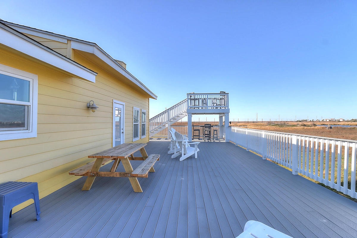 Expansive waterfront deck with picnic table and Adirondack chairs overlooking open marshland under clear blue skies.