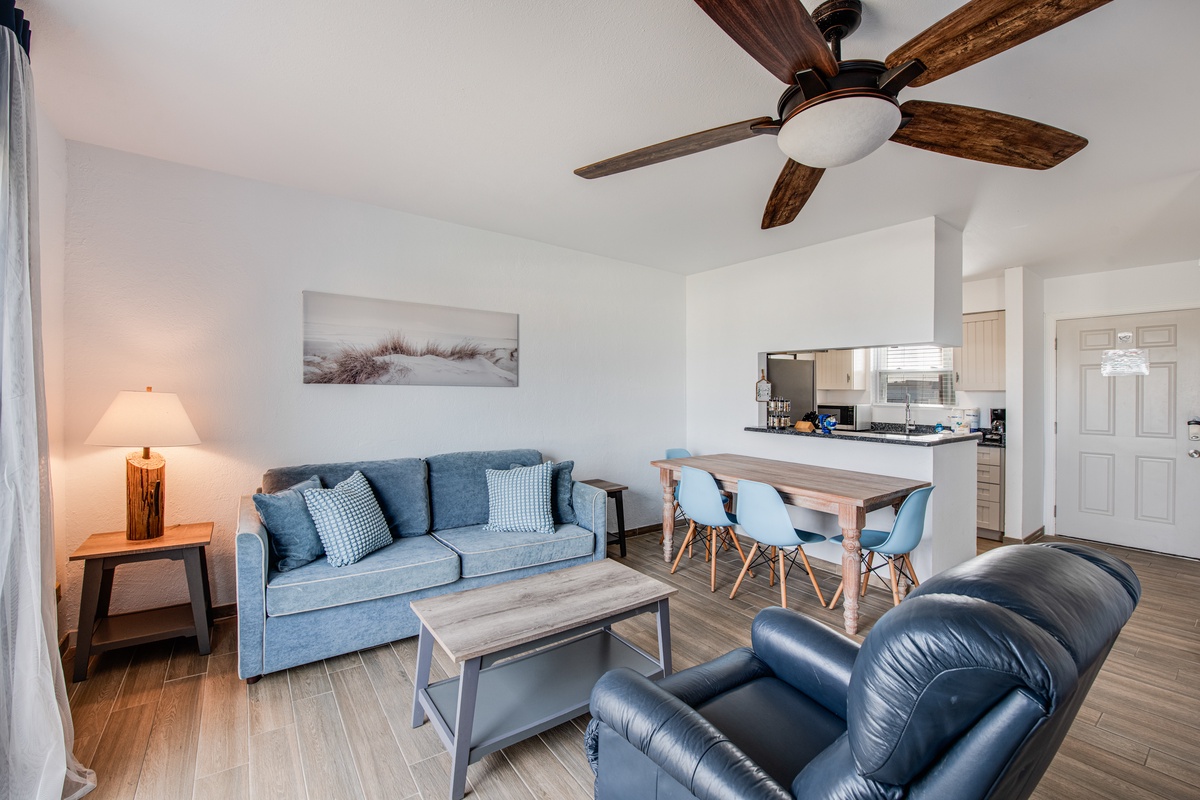 Relax in your coastal-styled living space with blue accents, wood-beam ceiling fan, and modern open layout connecting to the kitchen.