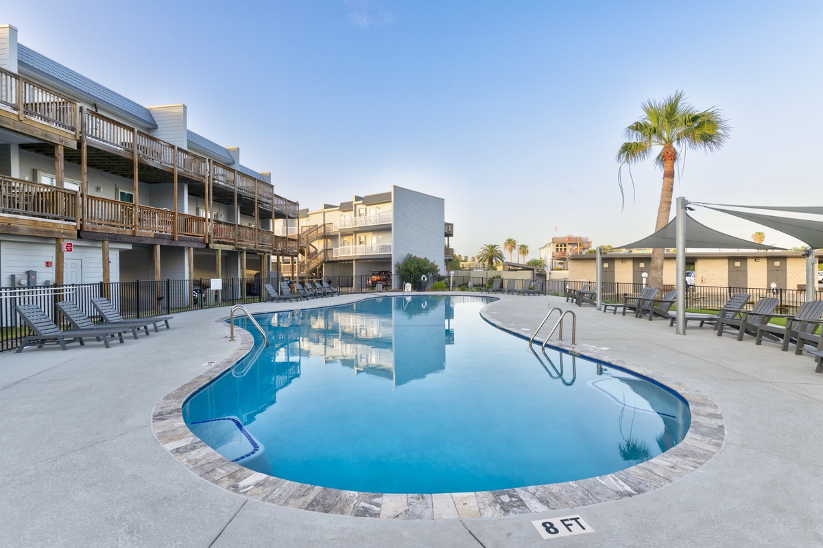 A sparkling pool with shaded seating awaits, surrounded by palm trees and modern buildings that frame your tropical escape under sunny skies.