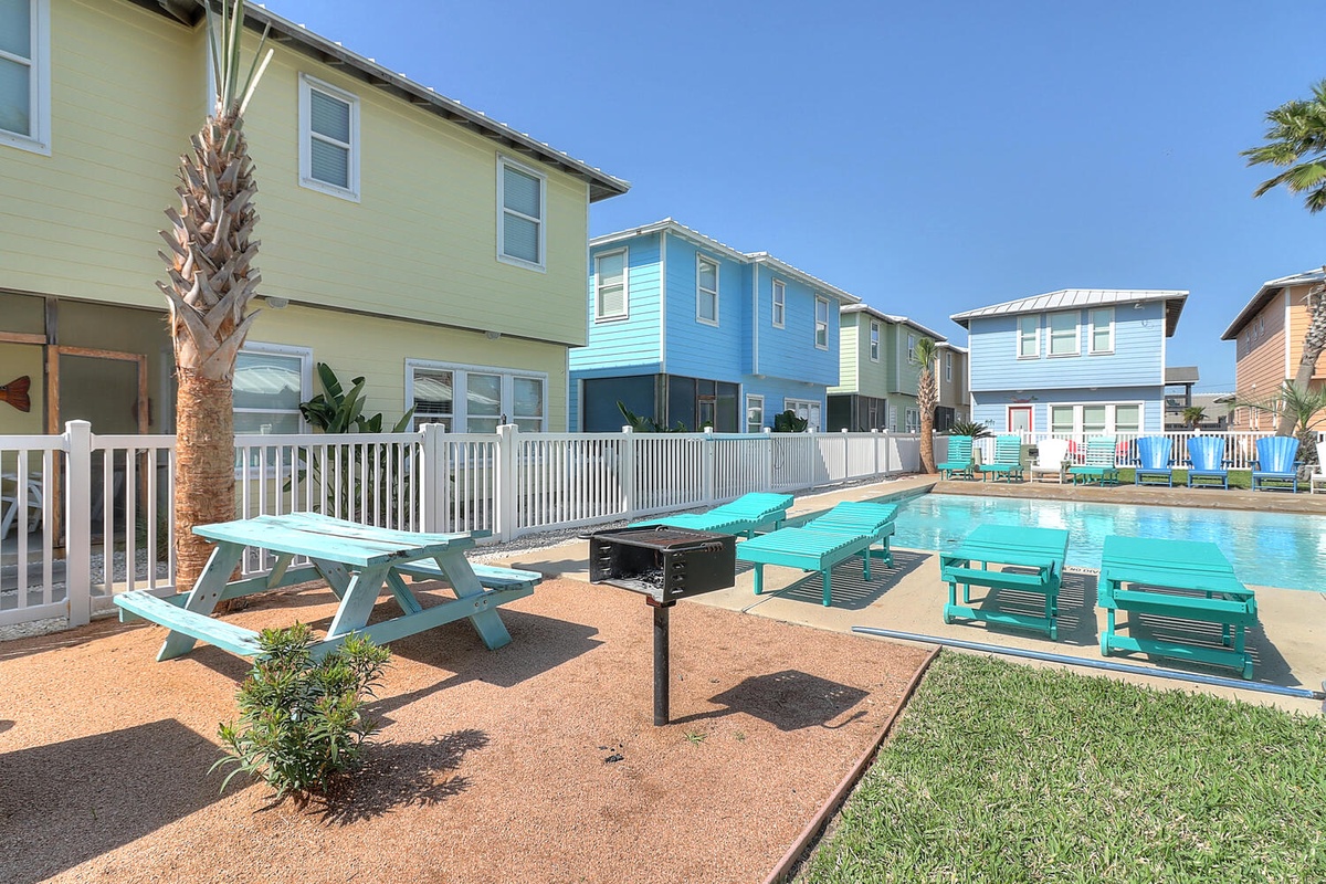 Colorful beach houses surround a sparkling swimming pool area with lounge chairs and outdoor dining space.
