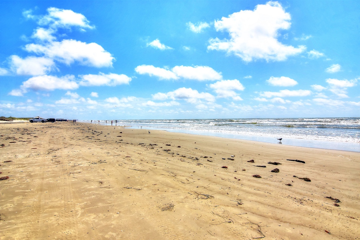 Wide sandy beach with gentle waves under bright blue skies dotted with fluffy white clouds, offering endless coastal beauty.