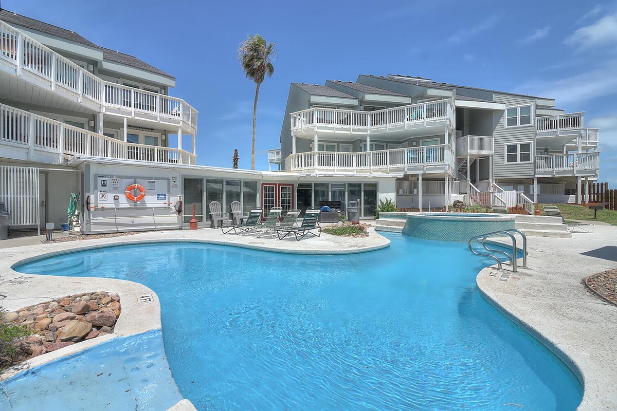 Coastal resort complex featuring multi-level apartment buildings with white balconies surrounding a curved swimming pool area.