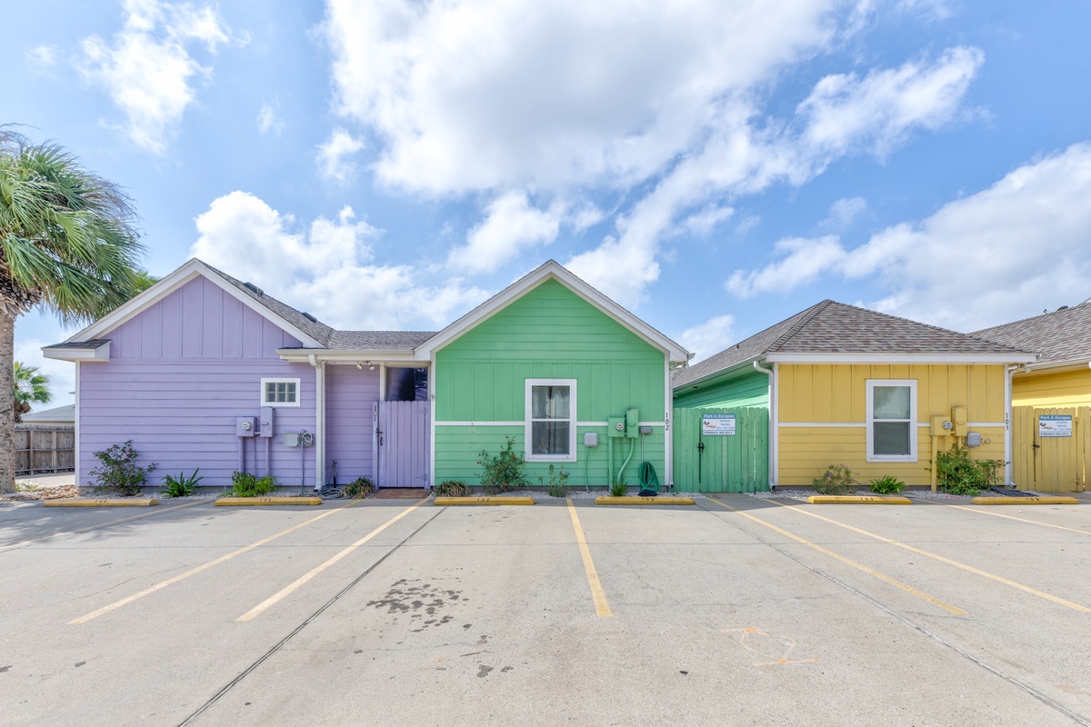 Charming colorful cottages featuring purple, green, and yellow exteriors with dedicated parking spaces under a beautiful blue sky.
