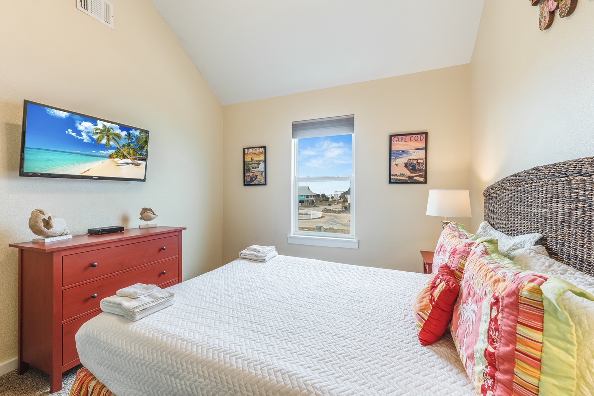 Your cozy bedroom features a comfortable bed, beach-themed TV, and colorful linens. Fresh towels await your arrival.