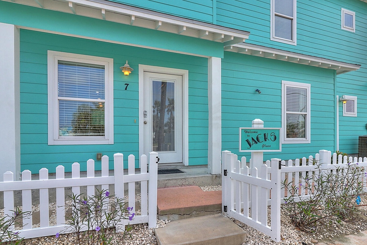 Charming turquoise beach cottage with white picket fence gate creates a welcoming coastal entrance to your waterfront retreat.