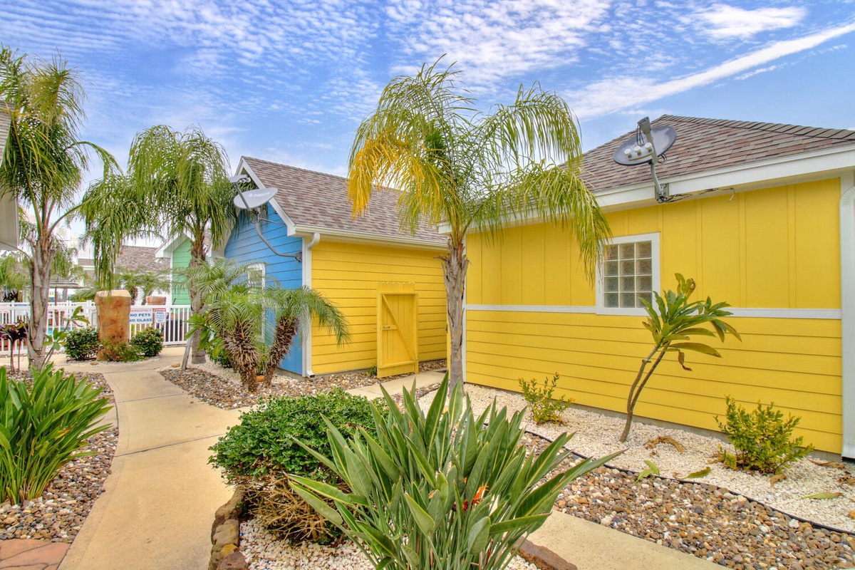 Vibrant yellow cottage welcomes guests with tropical palm trees and colorful neighboring units in a charming beachside community.