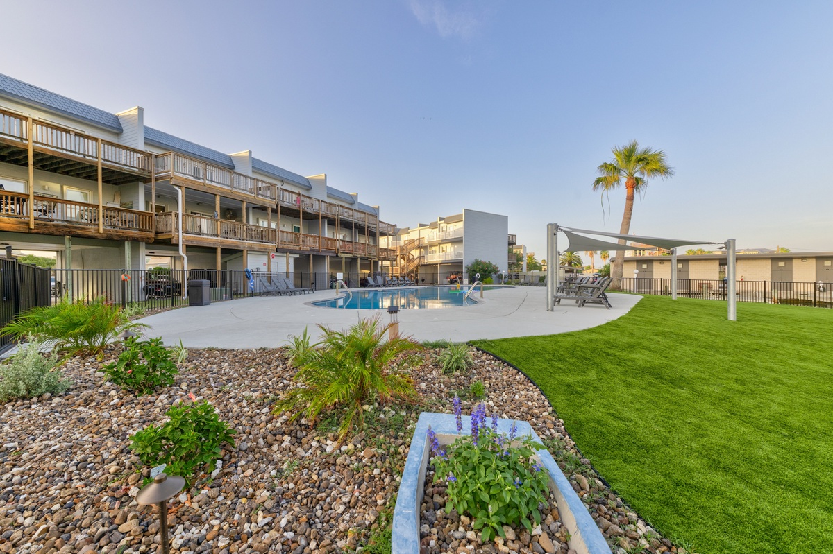 Multi-story property with balconies overlooking a sparkling pool, landscaped gardens with palm trees, and modern pergola seating areas. A spacious lawn provides additional outdoor space.