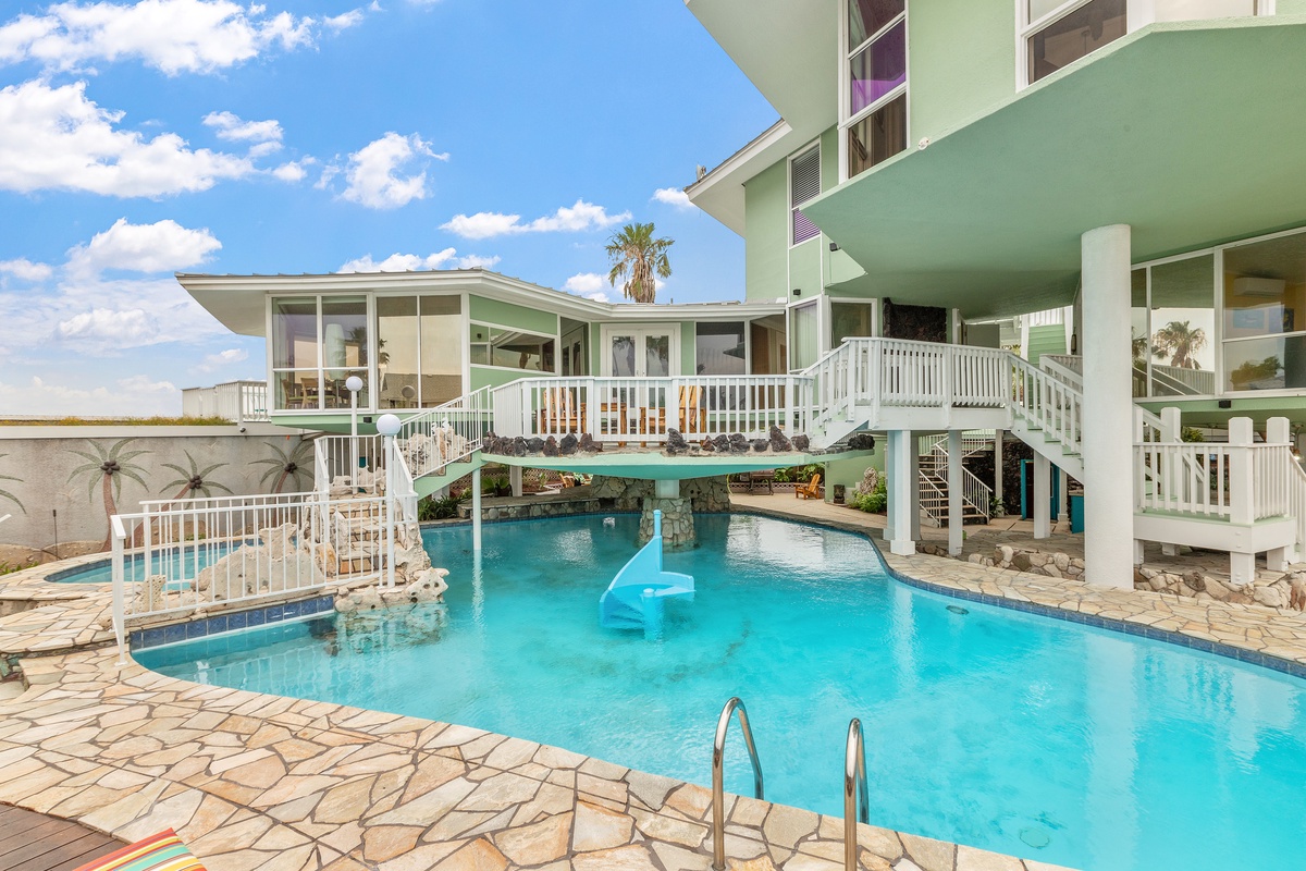 Modern beachfront property with stunning swimming pool featuring elevated walkways and crystal-clear turquoise waters under bright blue skies.
