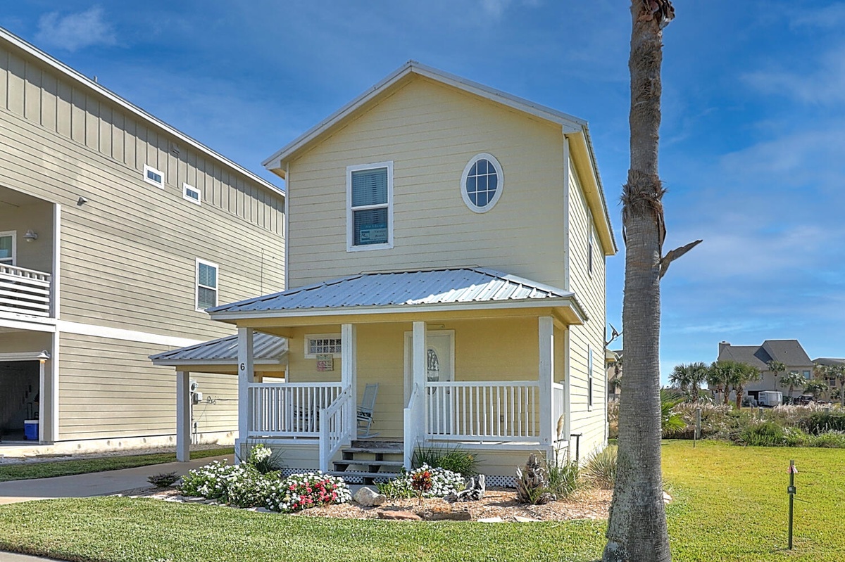 Charming coastal cottage featuring a welcoming front porch, tropical landscaping, and bright blue skies perfect for your beach getaway.