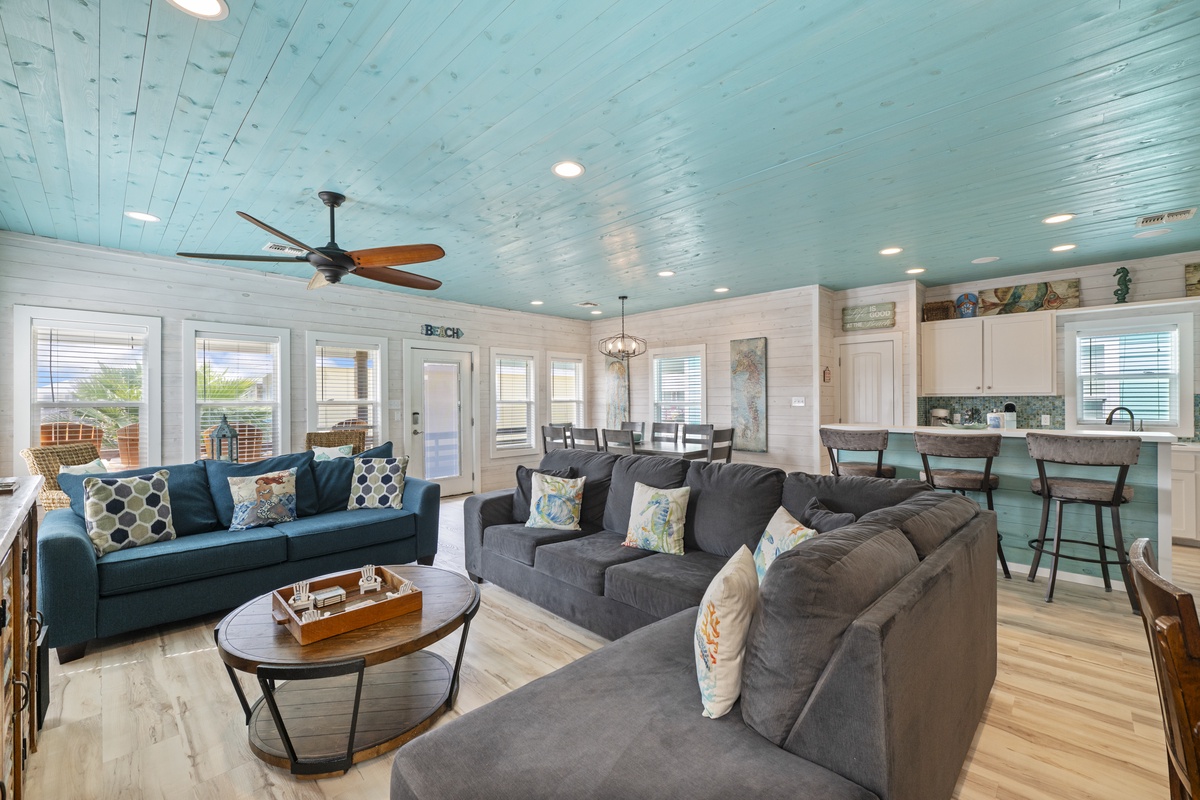 Relax in your spacious open-concept living area with coastal charm—a turquoise beadboard ceiling, ceiling fans, plush seating, and a bright kitchen with breakfast bar invite you to unwind in style.