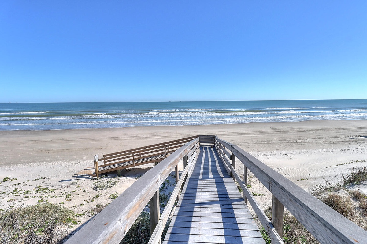 A wooden boardwalk leads to pristine sandy shores where gentle waves meet the coastline under a clear blue sky, offering direct beach access for relaxing days by the sea.