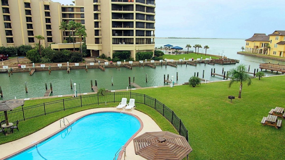 Waterfront resort with swimming pool overlooking marina and coastal waters, surrounded by palm trees and green lawns.