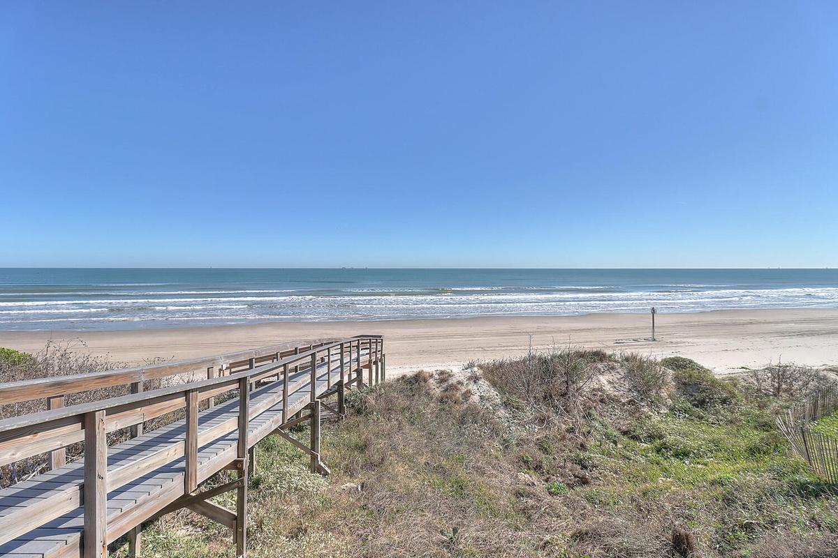Expansive oceanfront setting with private beach access via wooden walkway, stretching across pristine sand under brilliant blue skies.