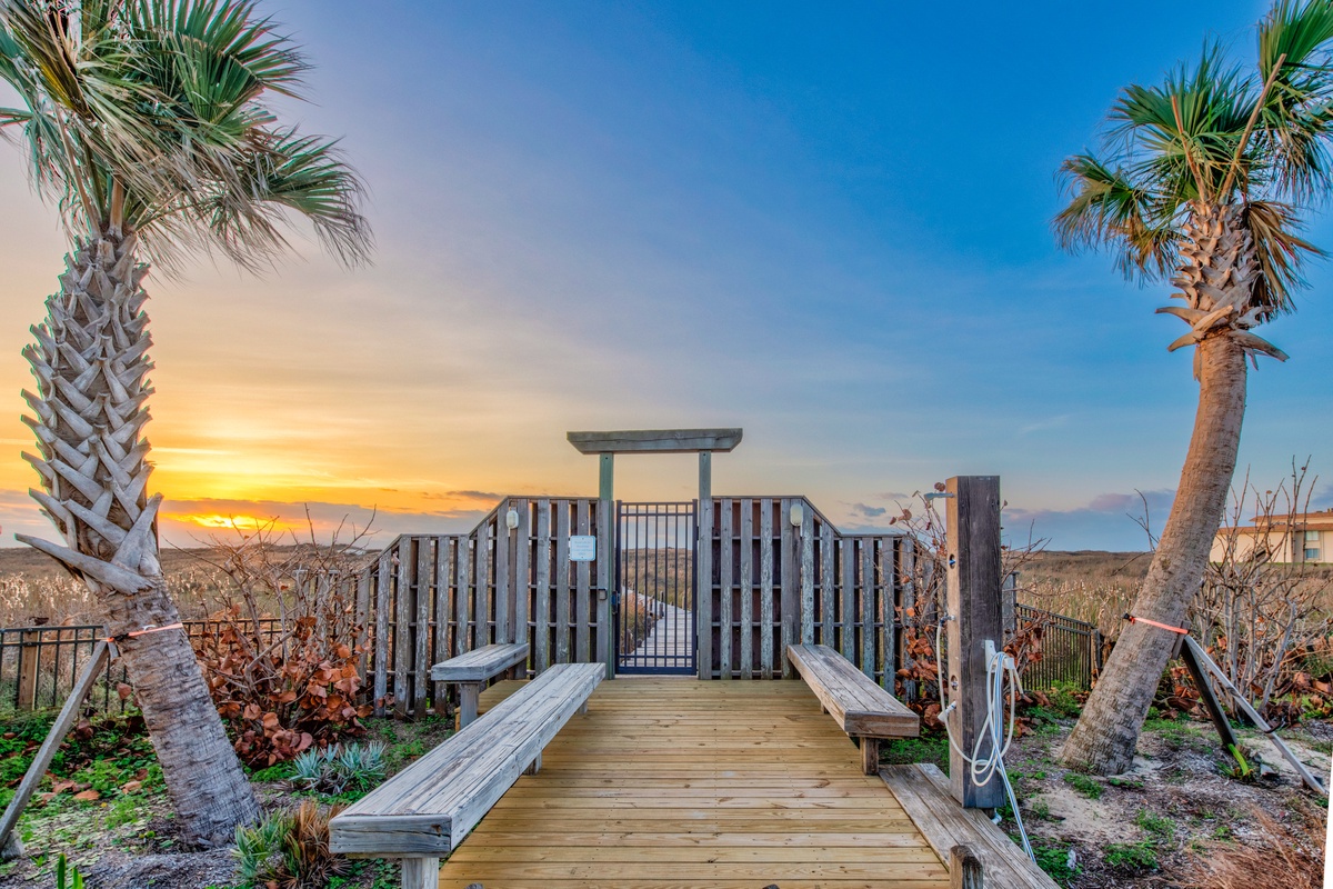 Wooden boardwalk leads through tropical palms toward the beach, with golden sunset painting the sky in warm coastal hues.