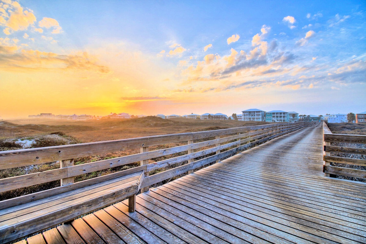 Wooden boardwalk stretches across coastal dunes toward residential homes beneath a stunning golden sunset sky.