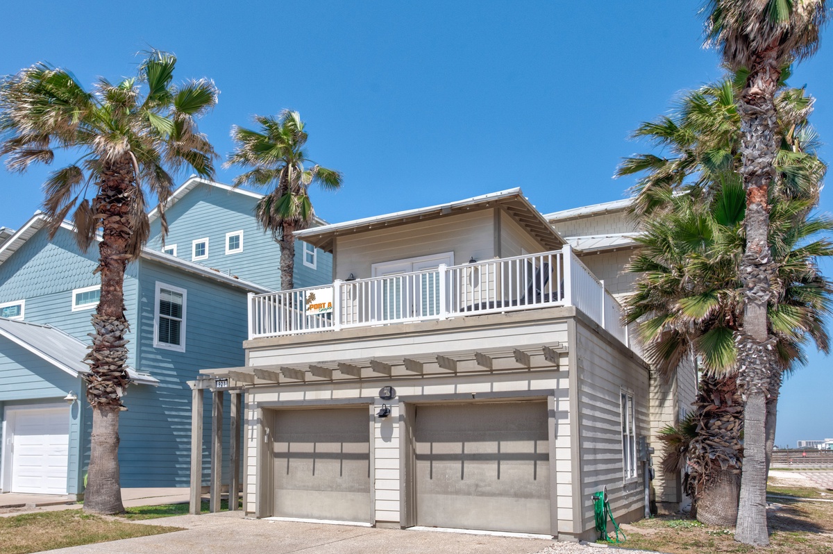 Modern coastal home featuring palm-lined tropical setting with convenient garage parking and upper-level balcony under brilliant blue skies.