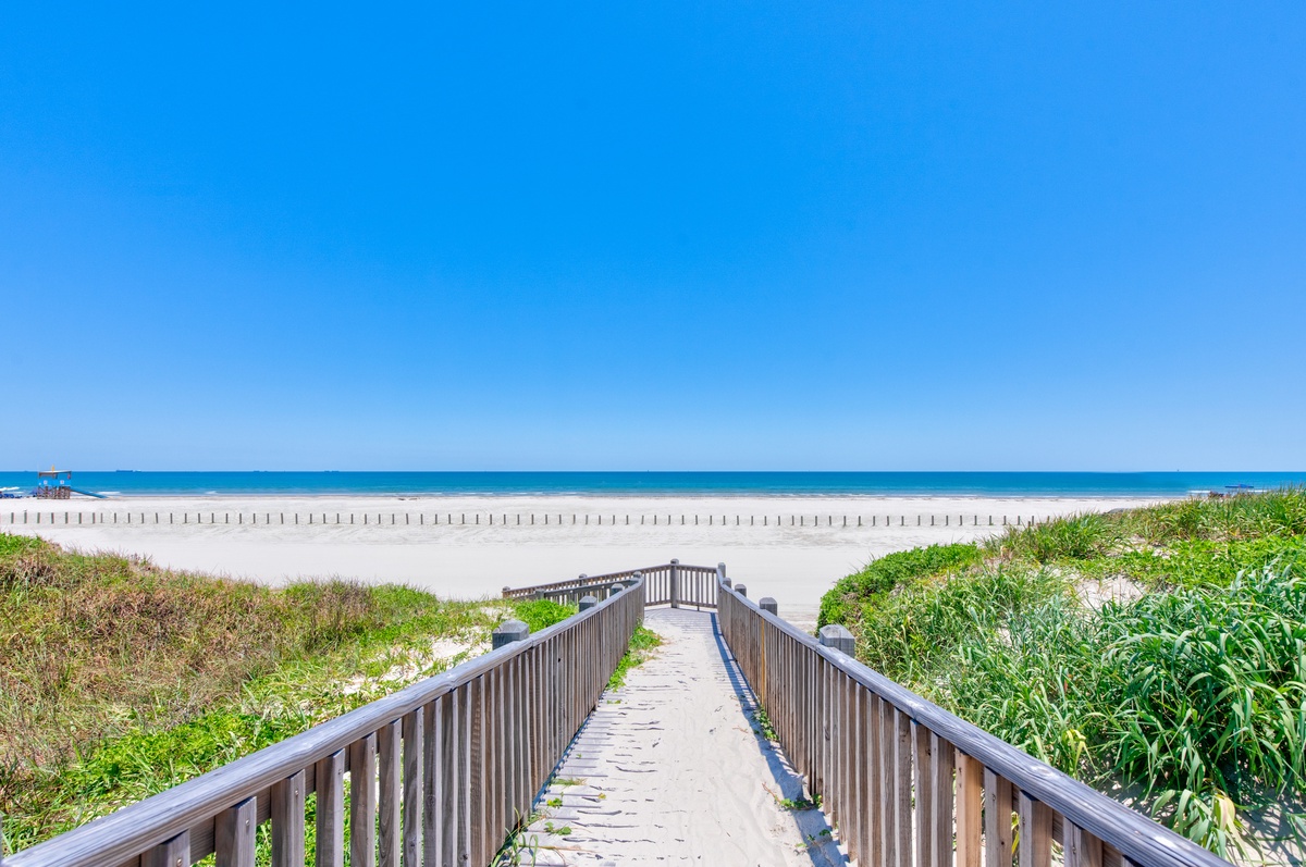 Wooden boardwalk leading to pristine white sand beach with crystal-clear turquoise waters under endless blue skies.